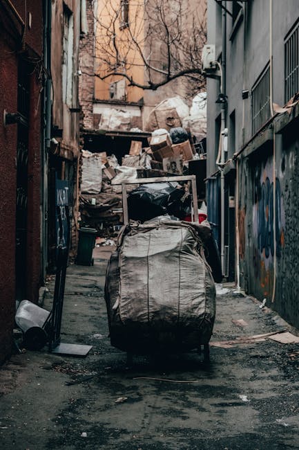 A narrow urban alleyway cluttered with discarded waste and debris, featuring a large, black, wheeled refuse bag in the foreground, which appears to be filled with rubbish. Behind it, there are multiple stacked cardboard boxes, plastic bags, and miscellaneous waste materials piled high against a weathered brick wall at the end of the alley. The alley is flanked by tall buildings with a mix of metal and brick exteriors; one side has a graffiti-covered metal door, while the other side shows barred windows and a grey wall with visible utility cables. Sparse bare tree branches extend over the scene, indicating an outdoor setting in colder months. The ground surface is cracked asphalt with dark stains, and the scene has a dull, subdued lighting typical of overcast weather. The image subtly reflects the context of independent rubbish collection or on-site waste handling, aligning with private rubbish removal services such as Waste Clearance Hounslow.