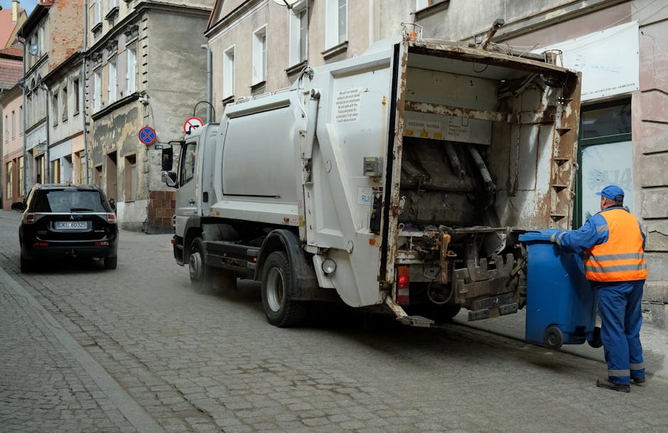 A waste collection vehicle from Waste Clearance Hounslow is parked on a narrow urban street, with its rear compaction mechanism open, revealing a rusted metal surface and internal components used for collecting and compacting rubbish. The vehicle is white with minor signs of wear and rust around the rear section. A worker dressed in a blue uniform and bright orange high-visibility vest, wearing a blue cap, is positioned to the right, lifting a blue wheeled bin towards the truck's hopper for on-site waste disposal. The street features cobblestone paving and lined with aged, multi-storey buildings that have European-style facades, some showing peeling paint and weathered surfaces. Parked cars, including a dark SUV, are visible along the curbside. There is a clear natural light illuminating the scene, and signage indicating parking restrictions can be seen attached to the buildings. This scene exemplifies independent waste collection services suitable for addressing access issues in narrow streets, such as those encountered in Hounslow, facilitating private disposal and rubbish clearance outside the scope of local authority collection.