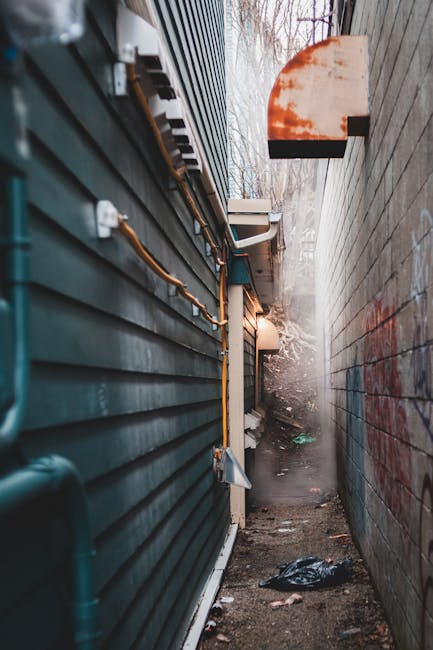 A narrow urban alleyway cluttered with discarded waste and debris, featuring a large, black, wheeled refuse bag in the foreground, which appears to be filled with rubbish. Behind it, there are multiple stacked cardboard boxes, plastic bags, and miscellaneous waste materials piled high against a weathered brick wall at the end of the alley. The alley is flanked by tall buildings with a mix of metal and brick exteriors; one side has a graffiti-covered metal door, while the other side shows barred windows and a grey wall with visible utility cables. Sparse bare tree branches extend over the scene, indicating an outdoor setting in colder months. The ground surface is cracked asphalt with dark stains, and the scene has a dull, subdued lighting typical of overcast weather. The image subtly reflects the context of independent rubbish collection or on-site waste handling, aligning with private rubbish removal services such as Waste Clearance Hounslow.