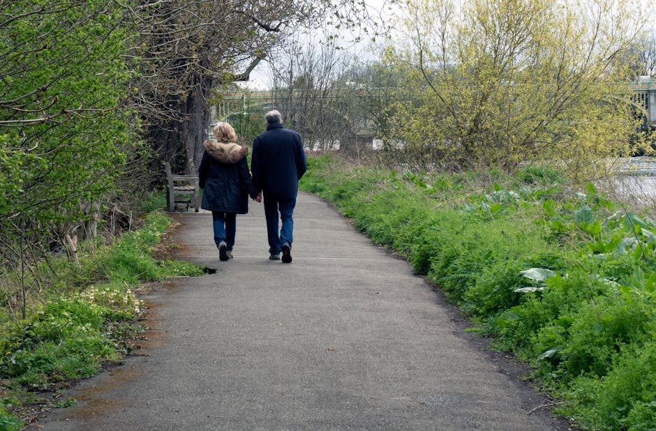 A couple is walking along a paved pathway in a park or green space during early spring or late autumn, as indicated by the budding trees and presence of some leaves. The woman is wearing a dark coat with a fur-lined hood, and the man is dressed in a darker jacket and blue jeans. They are holding hands as they walk away from the camera, with their backs visible. To the left of the pathway, there is a wooden bench situated beneath a large, leafless tree with textured bark, suggesting it is an older tree. The surrounding environment includes lush green grass and various small plants along the edges of the path, with some patches of bare soil. On the right side, the foliage is dense, with leafy bushes and small trees, some showing new growth. In the background, more trees and a hint of a body of water or river are visible, along with distant structures that are partially obscured by branches. The scene appears calm and natural, emphasizing the peaceful environment often associated with private or independent outdoor outings that might relate to waste clearance or outdoor maintenance loosely connected to rubbish removal services.