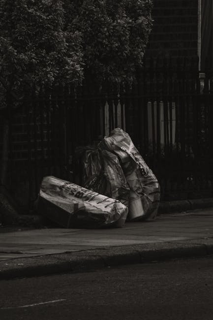 Two large black garbage bags filled with waste are positioned on a pavement near a curb, leaning against a black metal fence. The bags appear to be made of thick plastic and are tightly knotted at the top. One of the bags has some printed text visible on it, though it is not fully readable. Behind the fence, there are dense bushes or small trees with dark foliage, providing a backdrop of greenery. The environment appears to be an outdoor urban setting, possibly a residential area. The scene is captured in natural light, with shadows cast by nearby objects, highlighting the bags' crumpled texture and the rough surface of the pavement. The overall scene suggests the removal of household waste, which could be part of a private waste disposal or rubbish removal service, such as those offered by Waste Clearance Hounslow, designed to facilitate alternative waste handling outside local authority collections.