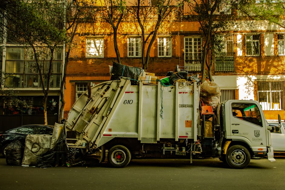 A white waste collection truck is parked on an urban street with a low curb, actively loaded with mixed rubbish materials. The truck’s large, enclosed body features a smooth, slightly curved surface with visible hinges and safety markings, and is labeled with the number 610. The materials inside include various bags, cardboard boxes, and bulky items, some of which are spilling slightly over the top edge. The surface of the rubbish varies in texture, with crumpled paper, plastic wrapping, and bulky construction debris, all combined into an uneven pile. The vehicle’s cab, positioned at the front, is compact with a side mirror visible on the door and a small window. Behind the truck, there are residential buildings with orange brick facades and numerous multi-pane windows reflecting daylight, along with a few leafy trees casting shadows across the street and building fronts. The scene is illuminated by natural sunlight, creating warm highlights and mild shadows, emphasizing the working aspect of independent rubbish removal typical of private waste disposal services, as occasionally operated by companies like Waste Clearance Hounslow.