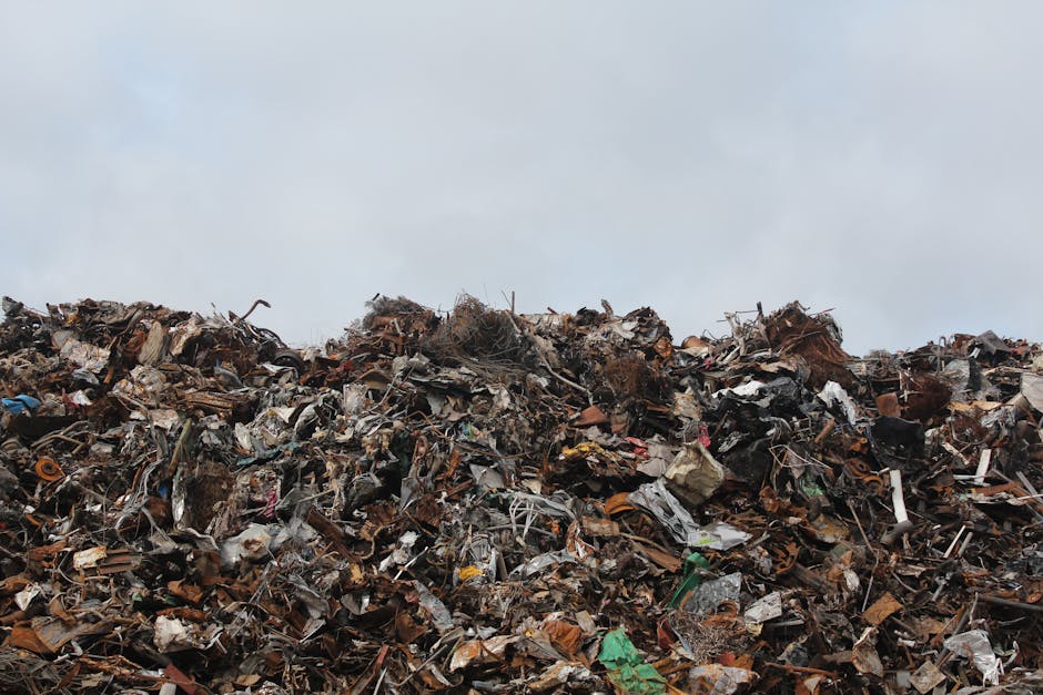 A large pile of mixed waste materials extends across the foreground, consisting of various types of trash including crumpled paper, plastic bottles, metal scraps, broken wood, and discarded packaging. The waste appears to be a combination of different textures and finishes, with some items shiny from plastics and metals, while others are dull and rusted. The pile is situated outdoors, likely on a paved or earth surface, with no visible containment or segregation. The background features an overcast sky with grey clouds, contributing to a muted, neutral atmosphere that emphasizes the scope of unmanaged rubbish. The scene reflects an environment that could be addressed through private waste removal or on-site clearance, aligning with services offered by Waste Clearance Hounslow for rubbish collection, which provides alternative disposal options outside local authority rubbish dumping or illegal dumping situations.
