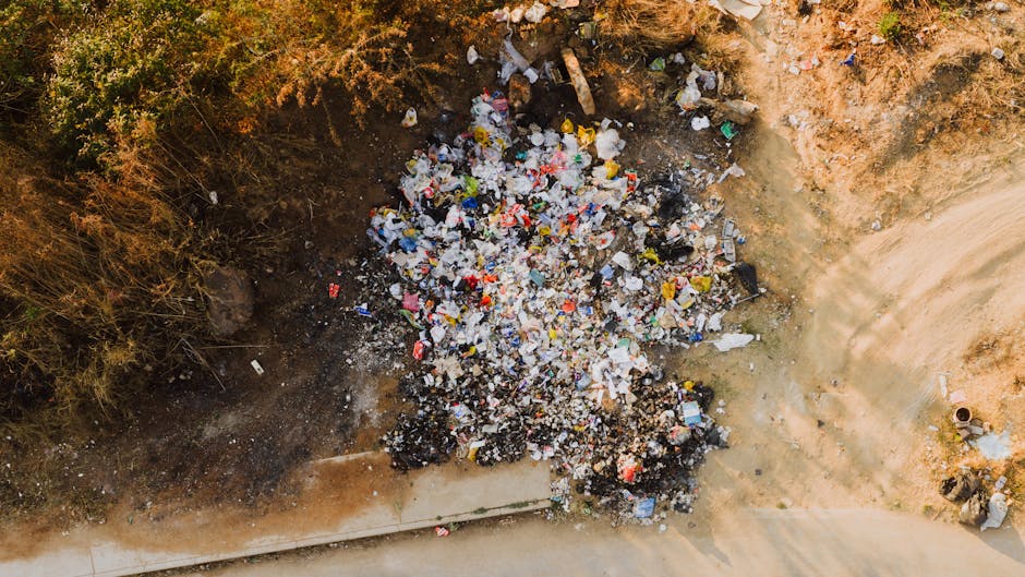 A large pile of mixed waste materials extends across the foreground, consisting of various types of trash including crumpled paper, plastic bottles, metal scraps, broken wood, and discarded packaging. The waste appears to be a combination of different textures and finishes, with some items shiny from plastics and metals, while others are dull and rusted. The pile is situated outdoors, likely on a paved or earth surface, with no visible containment or segregation. The background features an overcast sky with grey clouds, contributing to a muted, neutral atmosphere that emphasizes the scope of unmanaged rubbish. The scene reflects an environment that could be addressed through private waste removal or on-site clearance, aligning with services offered by Waste Clearance Hounslow for rubbish collection, which provides alternative disposal options outside local authority rubbish dumping or illegal dumping situations.