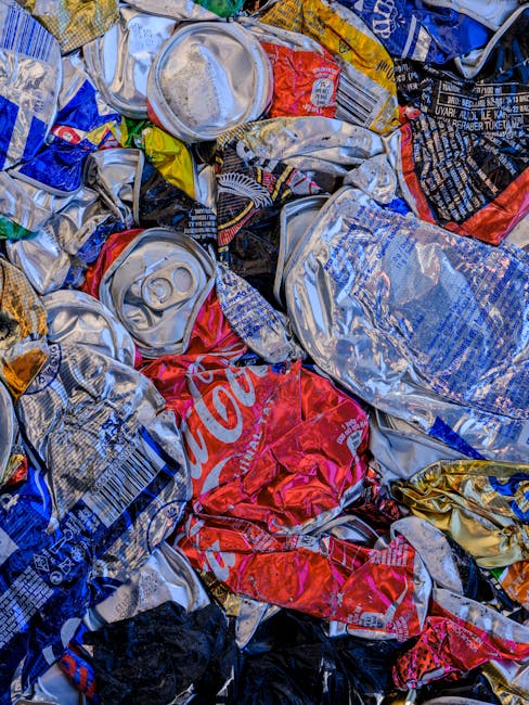 A close-up view of a pile of crushed aluminum cans and beverage containers scattered together, with various colors including silver, red, blue, yellow, and black. The cans exhibit crumpled and dented textures, with some partially flattened and others more rounded. Visible features include pull tabs, printed branding, and rippled surfaces, indicating different types of soft drink and energy drink cans. The background is filled with similar crushed cans, creating a cluttered scene typical of waste awaiting collection or disposal. Some cans display partially visible logos and labels, such as Coca-Cola, with parts of the branding distorted by crushing. The lighting appears natural, highlighting the reflective, metallic surface of the aluminum and emphasizing the textures of the crumpled metal objects. This image exemplifies waste ready for off-site rubbish collection, often handled by private waste clearance services like Waste Clearance Hounslow, as part of domestic or commercial rubbish removal processes.