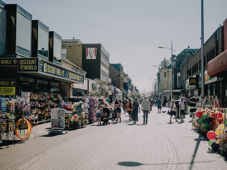 A busy pedestrian street in a town centre during the daytime, lined with various retail shops including a discount store, a fast-food outlet, and a clothing shop. The foreground shows numerous people walking, some with strollers or carts, engaging in shopping or casual strolling. On the left side, outdoor stall displays are visible, offering a variety of colorful items such as flowers, balloons, and toys, with some items stacked on the pavement and supported by metal racks. Building facades above the shops display signage and large windows; one shop has a prominent bright blue and yellow sign. To the right, additional retail units and a street lamp extend along the pavement, which appears clean and well-maintained. The overall atmosphere is lively with natural sunlight casting soft shadows, typical of an active shopping area. The scene visually relates to local, independent collection of retail waste or on-site clearance by waste management services, such as Waste Clearance Hounslow, illustrating typical urban rubbish management situations in a commercial district.