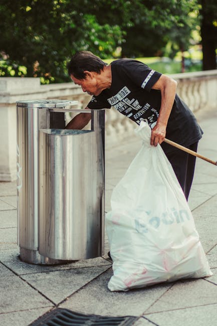 A middle-aged man with short dark hair wearing a black T-shirt with white text leans over a cylindrical metal waste bin on a paved sidewalk, actively disposing of rubbish. He holds a large white plastic bag filled with waste in his left hand, while his right hand is reaching into the bin to deposit trash. The man is standing outdoors in a park-like environment surrounded by lush green trees and foliage, which provide a shaded backdrop. The metal waste bin has a reflective, polished surface and an open top, positioned near a low stone wall or ledge on the left side of the image. The scene suggests an independent waste collection activity, typical of private rubbish removal services, emphasizing the importance of proper rubbish disposal on urban streets, supported by waste clearance specialists such as Waste Clearance Hounslow.