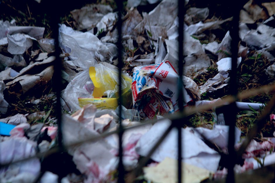 A close-up view of a pile of discarded trash and rubbish behind a black metal fence with vertical and horizontal bars. The collection includes crumpled paper, plastic bags, and food packaging, some of which are partially buried in dried leaves and dirt on the ground. The plastic bags are mainly white, with one yellow and another mainly white with red and blue printed details. The environment appears to be an outdoor area, possibly a neglected or unmanaged space. The lighting is natural but subdued, highlighting the chaos of the waste materials and the textured surfaces of the crumpled paper and plastic. This scene illustrates the importance of proper waste management and disposal, in line with independent rubbish collection services and facilities that handle hazardous or general waste effectively. Waste Clearance Hounslow provides solutions for clearing such unwanted debris in residential and commercial settings, supporting sustainable waste handling practices.
