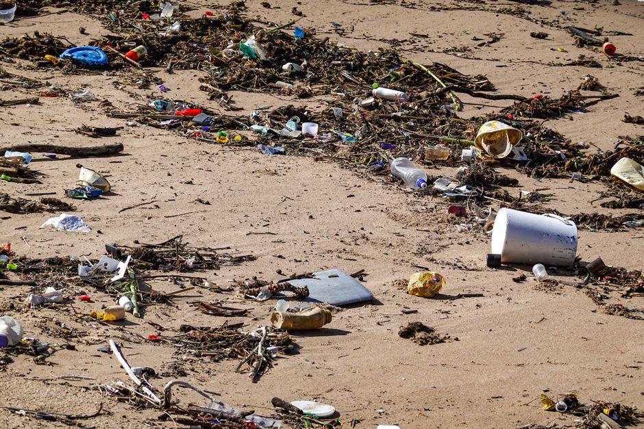A sandy beach scene showing a significant amount of litter scattered across the surface, including plastic bottles, cups, and containers, some partially buried in the sand while others are loosely strewn about. Various debris such as sticks, twigs, and small pieces of paper are also visible, mixed amongst the trash. The waste appears to be a combination of consumer waste and natural organic materials, creating an unkempt and polluted shoreline environment. The lighting suggests natural daylight, highlighting the contrast between the clean, sandy background and the tangible accumulation of discarded refuse. This image emphasizes the importance of proper waste disposal and the potential for private rubbish collection services, like those offered by Waste Clearance Hounslow, to mitigate environmental pollution and promote cleaner, healthier outdoor spaces.