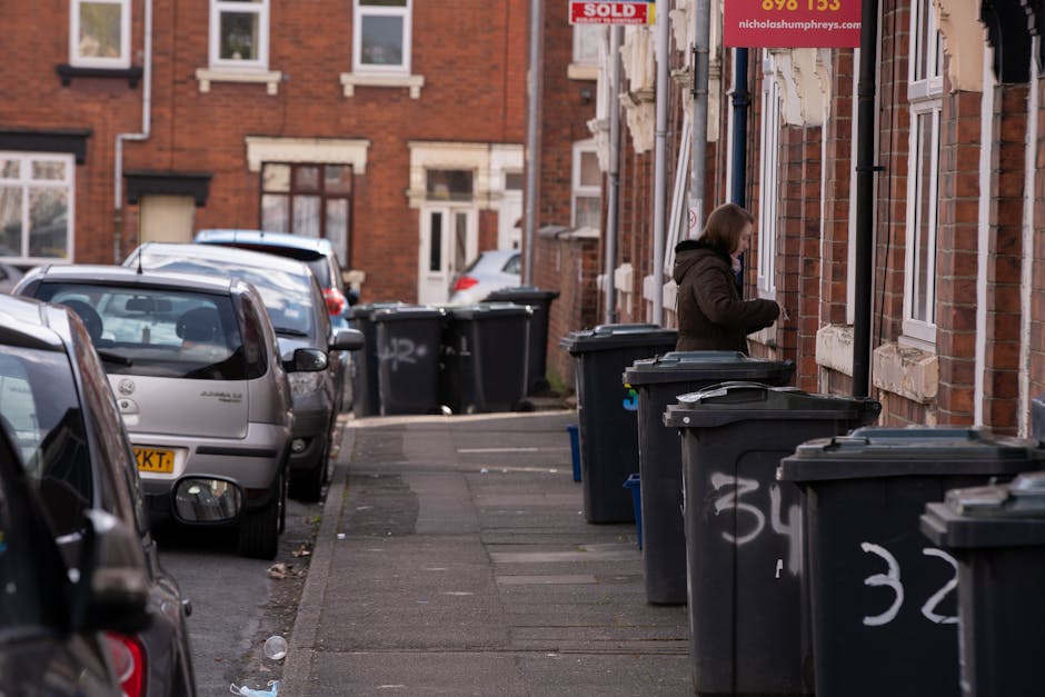 A row of black wheelie bins with closed lids are positioned along the sidewalk outside a residential building in a suburban area. The bins are made of plastic with a textured surface and are marked with white spray-painted numbers. Behind the bins, a woman dressed in a brown coat is seen standing near the brick exterior wall of the building, which features multiple windows with white frames and beige stone sills. The pavement and adjacent street are lined with parked cars in various shades of gray and silver, with some cars partially visible in the foreground. The environment appears to be in late afternoon lighting, casting soft shadows on the ground. A red and white property sign indicating 'sold' is visible above the woman, attached to the building façade, which has a reddish-brown brick finish. The scene subtly relates to waste management and private rubbish collection services, exemplified by the presence of the neatly arranged waste bins awaiting pickup, consistent with alternative rubbish clearance solutions offered by Waste Clearance Hounslow in the context of same-day rubbish clearance delays and solutions in Hounslow.