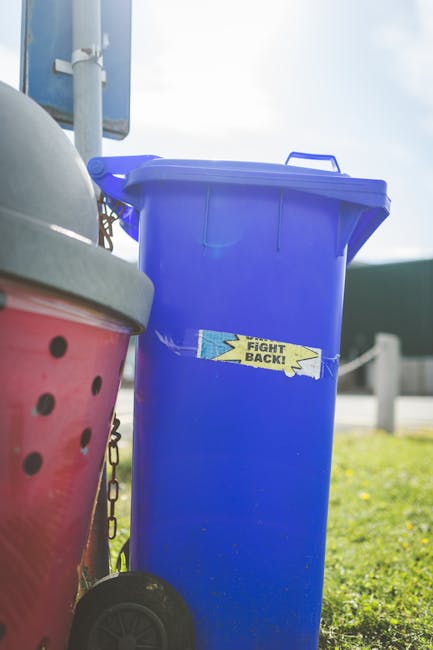 A tall, blue plastic rubbish bin with a closed lid positioned outdoors on a grassy area, next to a red wheeled bin with a partially visible lid. The blue bin has a slightly textured surface and a small, rectangular label with a yellow and blue design that reads 'FIGHT BACK!'. In the background, there is a metal pole, a blurry road with a barrier, and a bright sky suggesting daytime. The setting appears to be a typical outdoor environment where private rubbish collection or on-site waste disposal might occur, with the bins placed on a paved or grassy surface near a roadside or driveway. The scene is well-lit, with natural sunlight highlighting the bins' vibrant colors and creating a clean, professional impression related to waste clearance services focused on efficient rubbish removal.