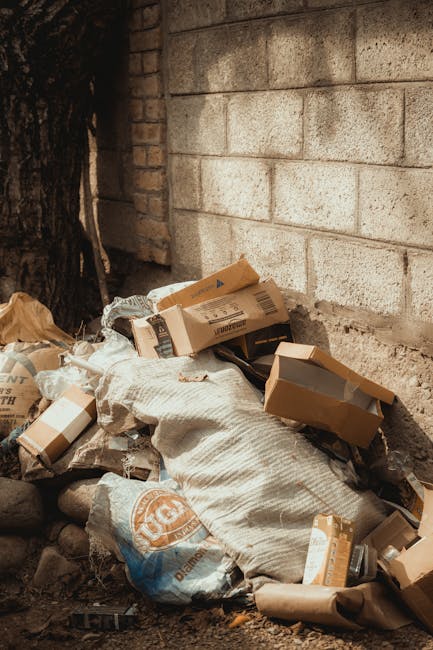 A pile of mixed rubbish including flattened cardboard boxes, plastic bags, and paper waste is accumulated against a brick and concrete wall, situated outdoors on a gravel surface. The cardboard boxes, some partially opened, are in various shades of brown and gray, with different sizes and textures, showing typical corrugated and smooth surfaces. A large, beige sack made of woven material is also visible among the debris, partially covering some of the boxes. The surrounding environment suggests an outdoor area, possibly a back alley or side yard, with minimal natural light illuminating the scene. The waste appears to be left for private collection or rubbish removal services such as Waste Clearance Hounslow, reflecting typical end-of-tenancy or domestic clearance scenarios. A sturdy tree trunk and rough brick wall are seen in the background, providing context for an informal rubbish storage spot before disposal or collection, relevant to alternative waste handling methods outside local authority bins.